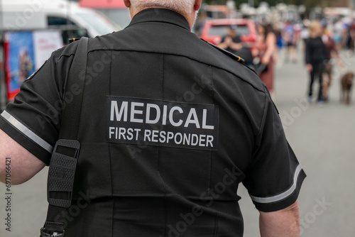 A close-up of a Caucasian male emergency health medical first responder or paramedic wearing a black uniform with grey letters. The ambulance attendant is wearing a short-sleeve shirt with lettering.