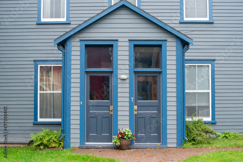 Two exterior half glass doors to a duplex. The building's wall is grey colored with royal blue trim and a pot of flowers in front. There are four double hung windows with curtains and blinds hanging. 