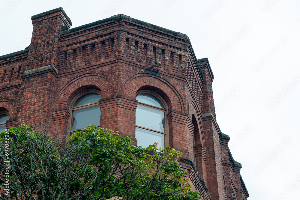 The exterior of a red or brown colored apartment building. The roof of the old structure is weathered with dark crevices in the brick. There's three closed glass windows with arched glass toppers. 