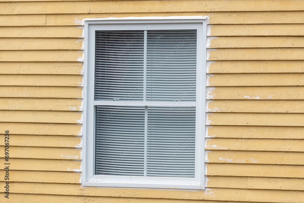 The exterior wall has a vibrant yellow wooden clapboard siding wall with a double-hung window. The wide colonial trim around the window is white. There are small white blinds in the window. 