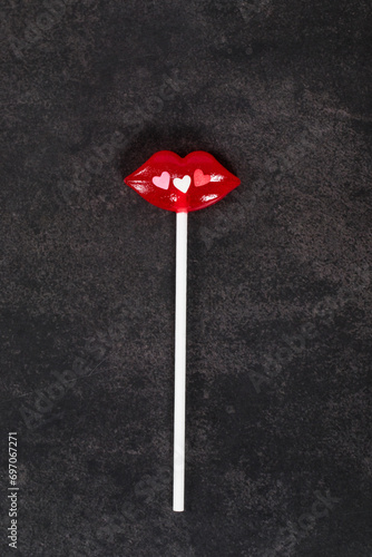 Red lips lollipop decorated with small colored sugar hearts. On a paper stick. Dark background. Top view. Valentine's day