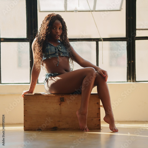 Girl with stunning figure in denim shorts poses near a large window while sitting on a wooden box. Beautiful young woman sitting in the studio with a large window during the day