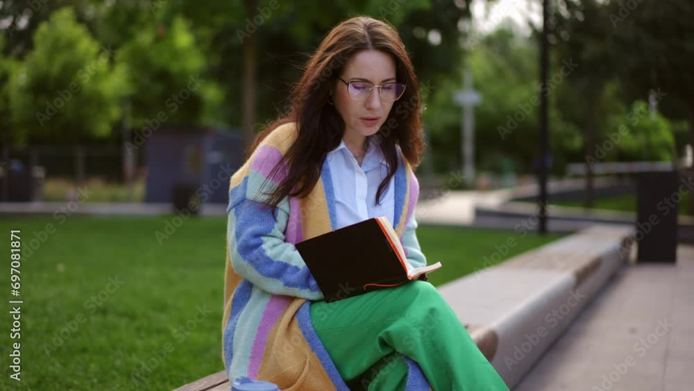 A young woman with long hair wearing glasses plans a work schedule, writing down her ideas with a pen in a notepad, sitting on a bench in a park. A young woman with long hair wearing glasses plans a work schedule, writing down her ideas with a pen in a notepad, sitting on a bench in a park.