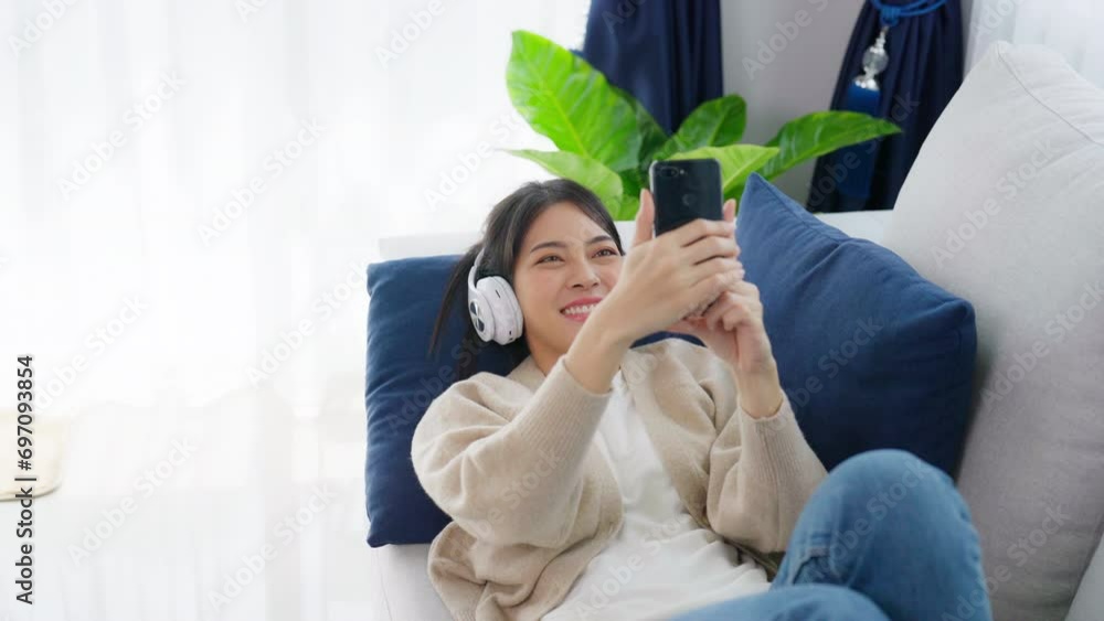 Happy young asian woman wearing headset and relax on comfortable couch at home texting messaging on smartphone, smiling girl use cell phone video call Happy young asian woman wearing headset and relax on comfortable couch at home texting messaging on smartphone, smiling girl use cell phone video call