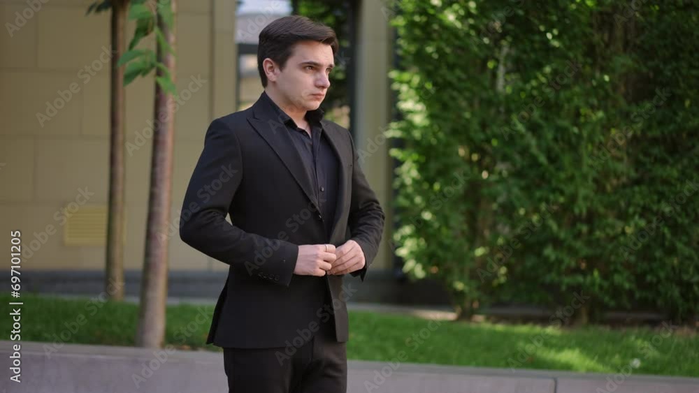 A young man in a black formal suit adjusts and buttons his jacket, while standing on the street against the backdrop of trees in the city A young man in a black formal suit adjusts and buttons his jacket, while standing on the street against the backdrop of trees in the city