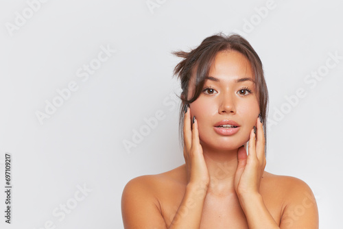 Young woman stands with naked shoulders over studio background pressing her hands to her face  skincare product concept  copy space