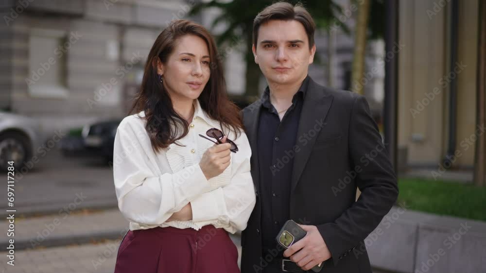 A businessman and a businesswoman pose for the camera together while standing on the street in the city. The man looks at the camera and the woman looks into the distance, then the woman looks at the A businessman and a businesswoman pose for the camera together while standing on the street in the city. The man looks at the camera and the woman looks into the distance, then the woman looks at the