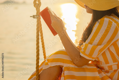 Lazy time. Asian Woman in yellow hat sitting on swing with book on a summer day. Sunset on a background a sea. Golden sunshine sea water surface. 