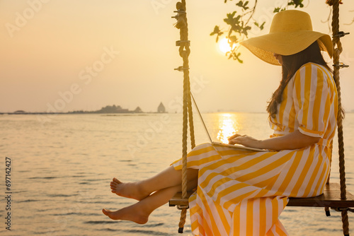 Happy traveler asian woman wearing long yellow dress and yellow hat using laptop while sitting on swing at sand beach. Beautiful sunset above sea or ocean, reflection of sun in the water.