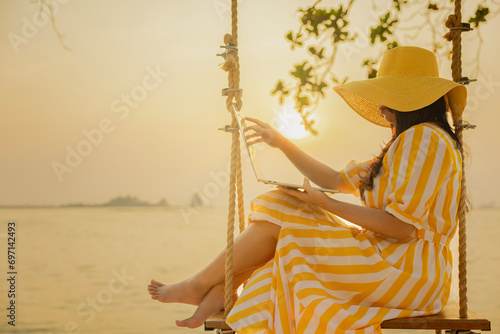Woman in yellow dress flying on wind while sitting on swing, working typing emails, browsing online on laptop, enjoying orange and golden sunset sky calmness tranquil relaxing sunlight summer mood.
