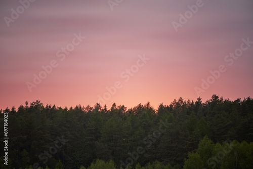 Evening Descends with a Colorful Display of Nature’s Beauty Over the Tranquil Woods. The Horizon Lights Up with Warm Colors as Dusk Falls Over a Peaceful Forest. © AlexGo