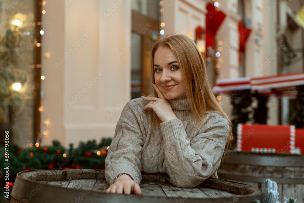 Attractive woman in sweater next to a table in a street cafe. Christmas decorations in the background. Cold season, walk in the city Attractive woman in sweater next to a table in a street cafe. Christmas decorations in the background. Cold season, walk in the city