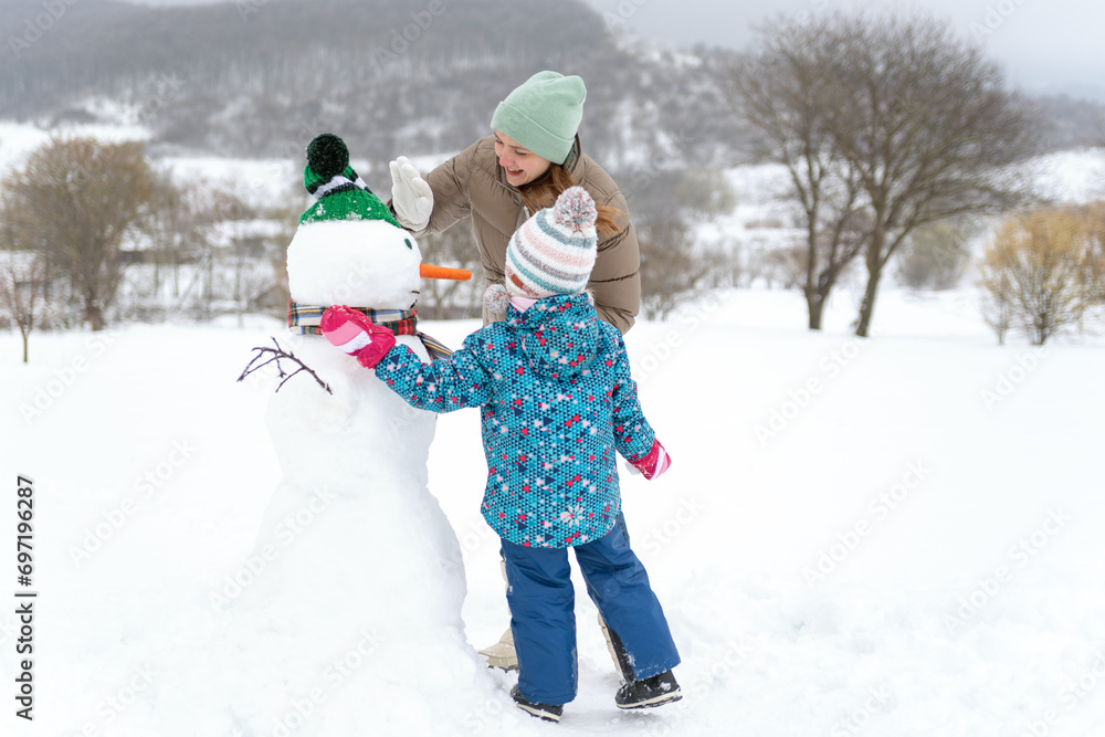 Mother and Toddler build large snowman with carrot nose. Kid during stroll in a snowy winter park. Mother and Toddler build large snowman with carrot nose. Kid during stroll in a snowy winter park.