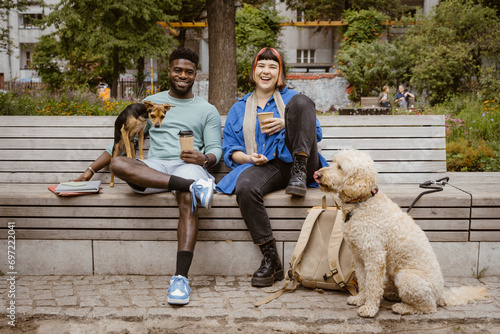 Happy young man and woman holding disposable coffee cups while sitting with dogs in park photo