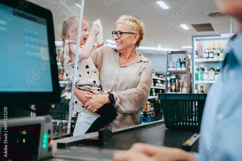 Happy senior woman carrying cheerful granddaughter holding bill at supermarket photo