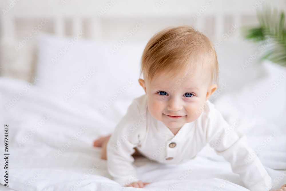 smiling baby 6 months old blond boy crawls on a white bed in a bright bedroom after sleeping in the morning in a cotton bodysuit, the concept of children's goods smiling baby 6 months old blond boy crawls on a white bed in a bright bedroom after sleeping in the morning in a cotton bodysuit, the concept of children's goods
