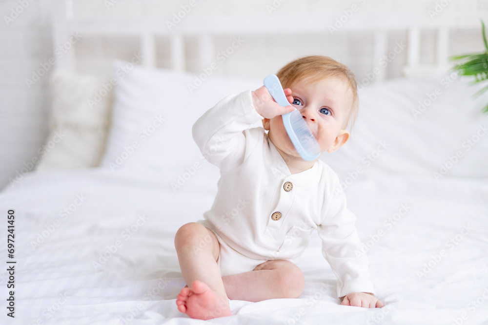 a 6-month-old blond boy is gnawing a comb on a white bed in a bright bedroom in a cotton bodysuit, a small child is teething - obrazy, fototapety, plakaty 