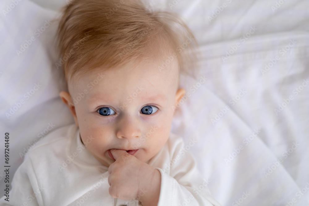 smiling baby 6 months old blond boy lies on a white bed in a bright bedroom and licks his finger in a cotton bodysuit, the concept of children's goods smiling baby 6 months old blond boy lies on a white bed in a bright bedroom and licks his finger in a cotton bodysuit, the concept of children's goods