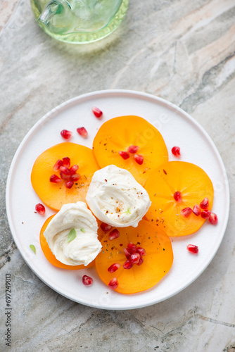 Sliced persimmon, mozzarella cheese and pomegranate on a white plate, vertical shot on a light-grey granite background, high angle view