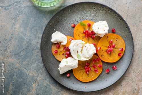 Plate with fresh persimmon slices, torn mozzarella and pomegranate seeds, high angle view on a beige and grey granite background, horizontal shot