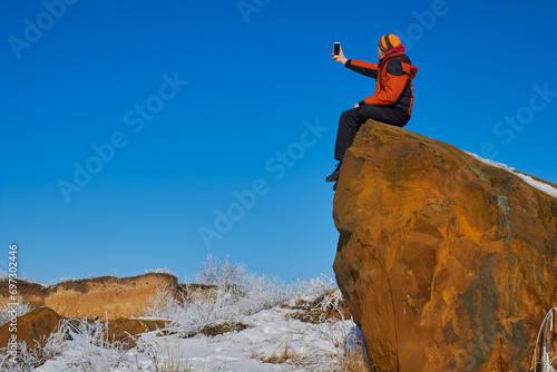 a tourist on a rock takes a selfie in winter, a man on a large sand stone in the mountains takes photos on a smartphone