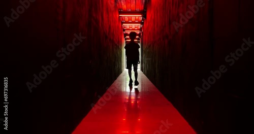Silhouetted young boy moves towards us in slow motion down dark corridor bathed in dramatic red light. His unsettling movements add to eerie atmosphere of scene, creating sense of mystery and suspense photo