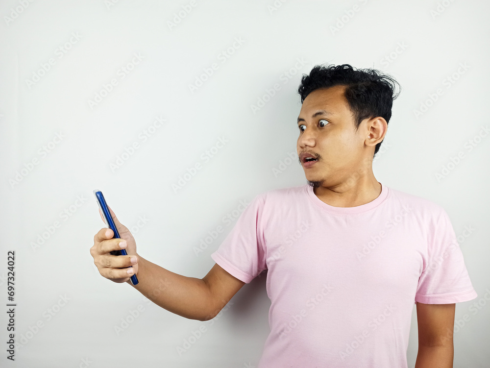 Shocked young Asian man in casual t-shirt looking at mobile phone screen, reacting to online news isolated on white background. People lifestyle concept. Advertising concept. Shocked young Asian man in casual t-shirt looking at mobile phone screen, reacting to online news isolated on white background. People lifestyle concept. Advertising concept.