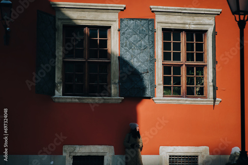 A young girl in a green down jacket and angora hat stands in profile near a terracotta-colored wall with windows and shutters in the rays of the setting sun with a dark shadow.Lviv, Armenian courtyard