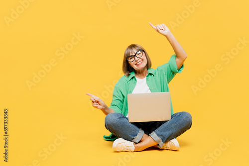 Full body elderly woman 50s years old wears green shirt glasses casual clothes sitting hold use work on laptop pc computer point aside on area isolated on plain yellow background. Lifestyle concept. photo