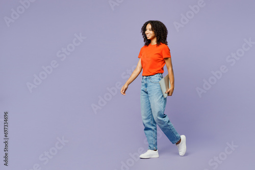 Full body side view little kid teen IT girl of African American ethnicity wear orange t-shirt hold closed laptop pc computer walk go isolated on plain purple background. Childhood lifestyle concept.