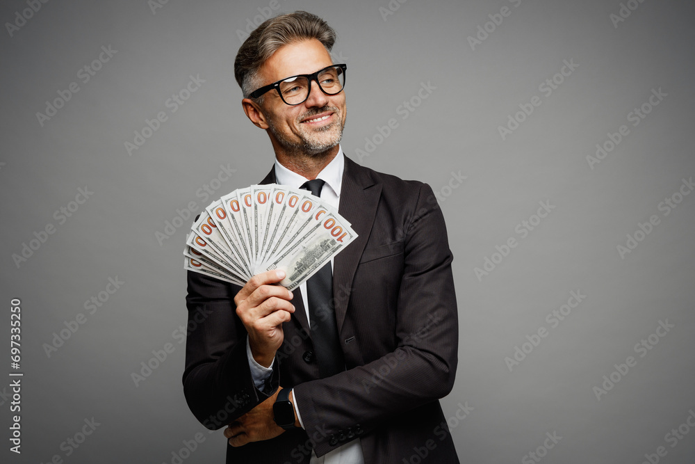 Adult rich happy employee business man corporate lawyer wear classic formal black suit shirt tie work in office hold fan of cash money in dollar banknotes look aside isolated on plain grey background. Adult rich happy employee business man corporate lawyer wear classic formal black suit shirt tie work in office hold fan of cash money in dollar banknotes look aside isolated on plain grey background.