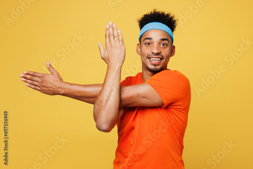 Young smiling happy fitness trainer sporty man sportsman wear orange t-shirt do exercise for hands look camera spend time in home gym isolated on plain yellow background Workout sport fit abs concept
