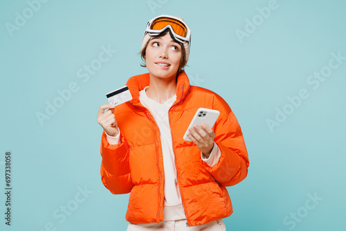 Skier young woman wear padded windbreaker jacket ski goggles mask hold credit bank card mobile cell phone travel rest going spend weekend winter season in mountains isolated on plain blue background.