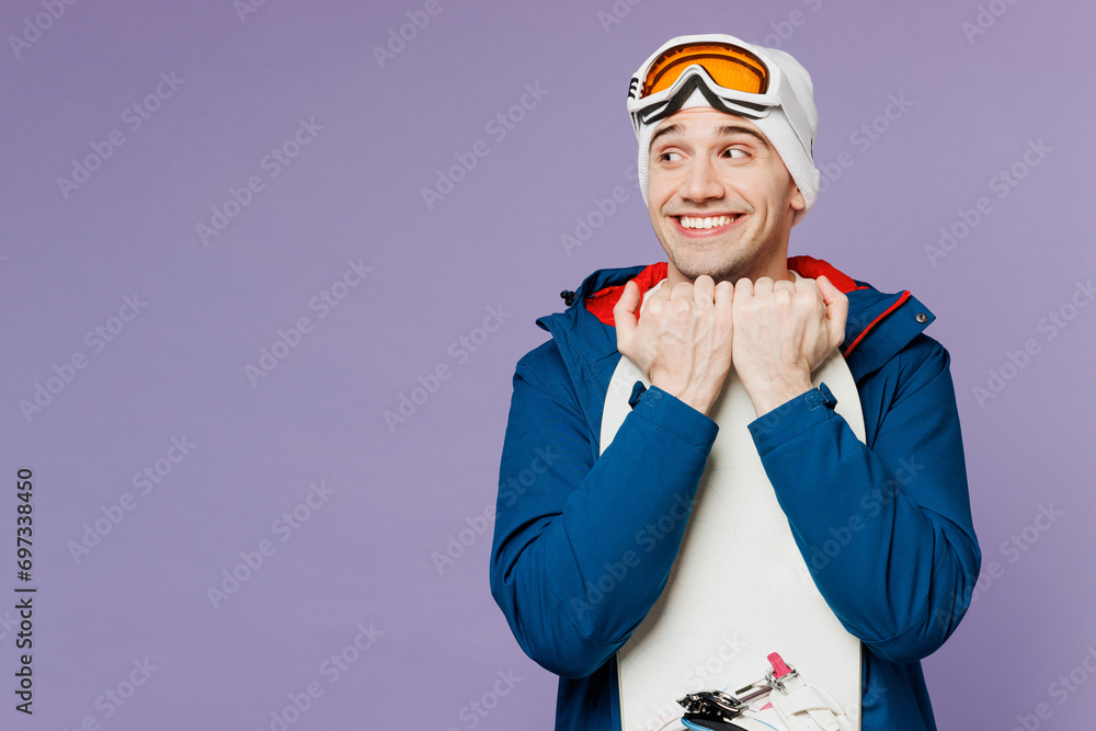 Obraz premium Amazed smiling happy man wear warm blue windbreaker jacket ski goggles mask hat hold snowboard look aside on area spend extreme weekend winter season in mountains isolated on plain purple background.