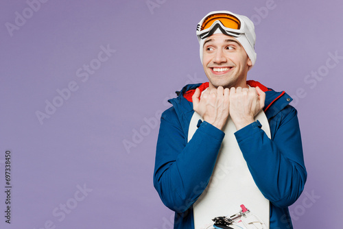 Amazed smiling happy man wear warm blue windbreaker jacket ski goggles mask hat hold snowboard look aside on area spend extreme weekend winter season in mountains isolated on plain purple background.