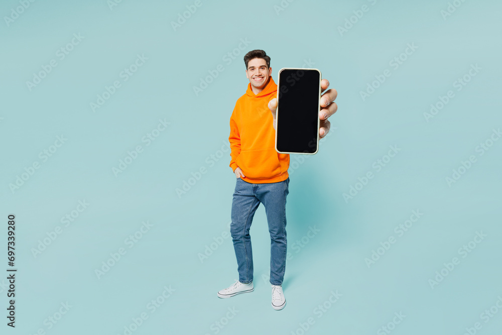 Full body young man he wears orange hoody casual clothes hold in hand use blank screen mobile cell phone isolated on plain pastel light blue cyan color background studio portrait. Lifestyle concept. - obrazy, fototapety, plakaty 