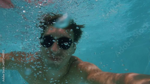 Funny man in sunglasses enjoys summer days, sunbathing swims in the pool and dives underwater filming himself with an action camera making a selfie video. POV video. photo