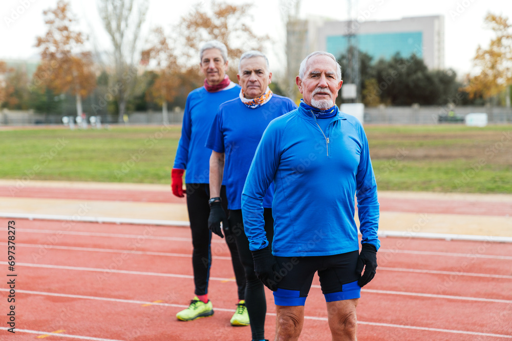 Three senior male runners in blue sportswear prepare for a track run, showcasing fitness and determination at an outdoor stadium.