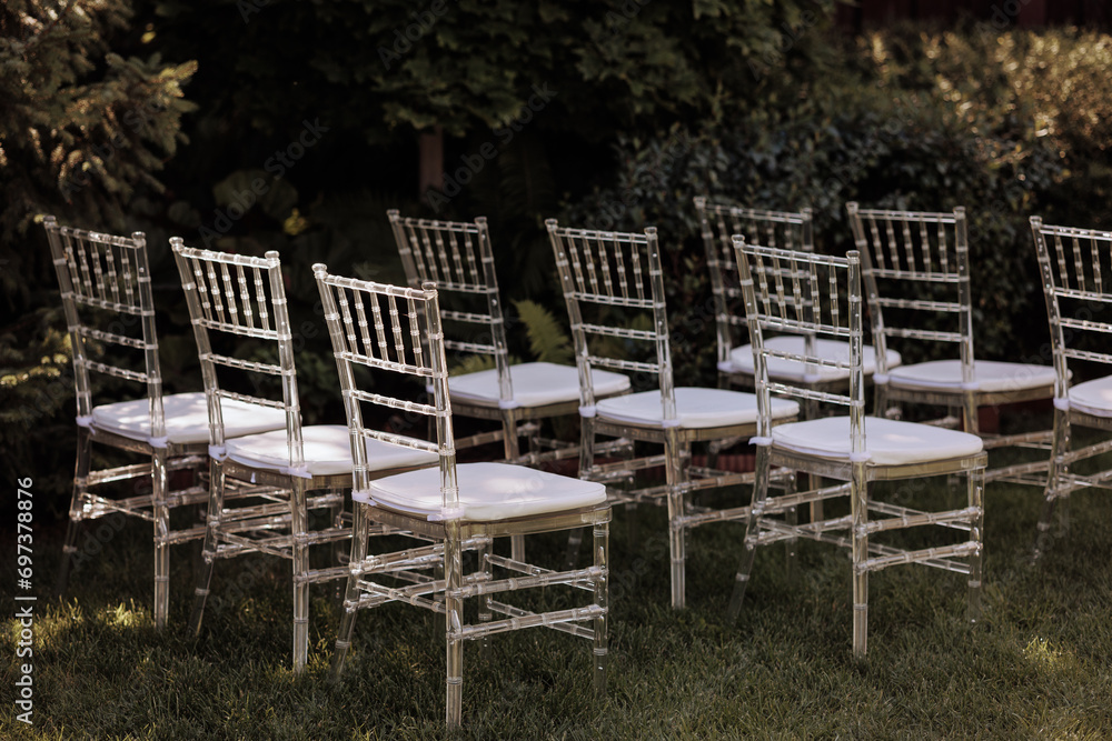 Row of elegant, transparent chairs with white cushions set outdoors in soft sunlight. Concept: outdoor events, modern design, or garden parties. Plenty of copy space.
