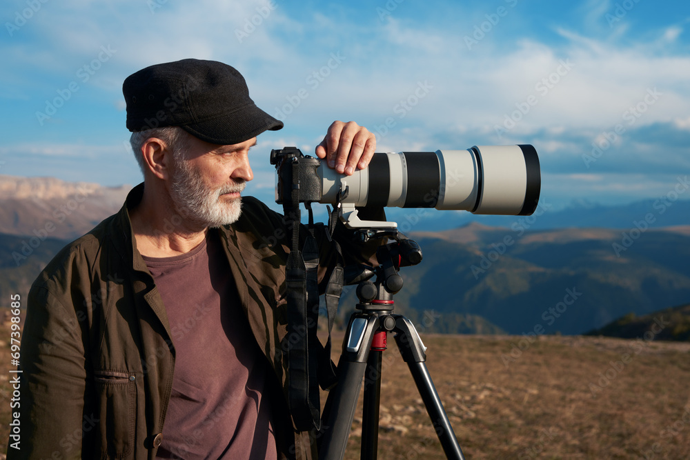 Photogaph (adult gray-haired man) with a camera with a long lens on a tripod on the background of mountainous terrain.