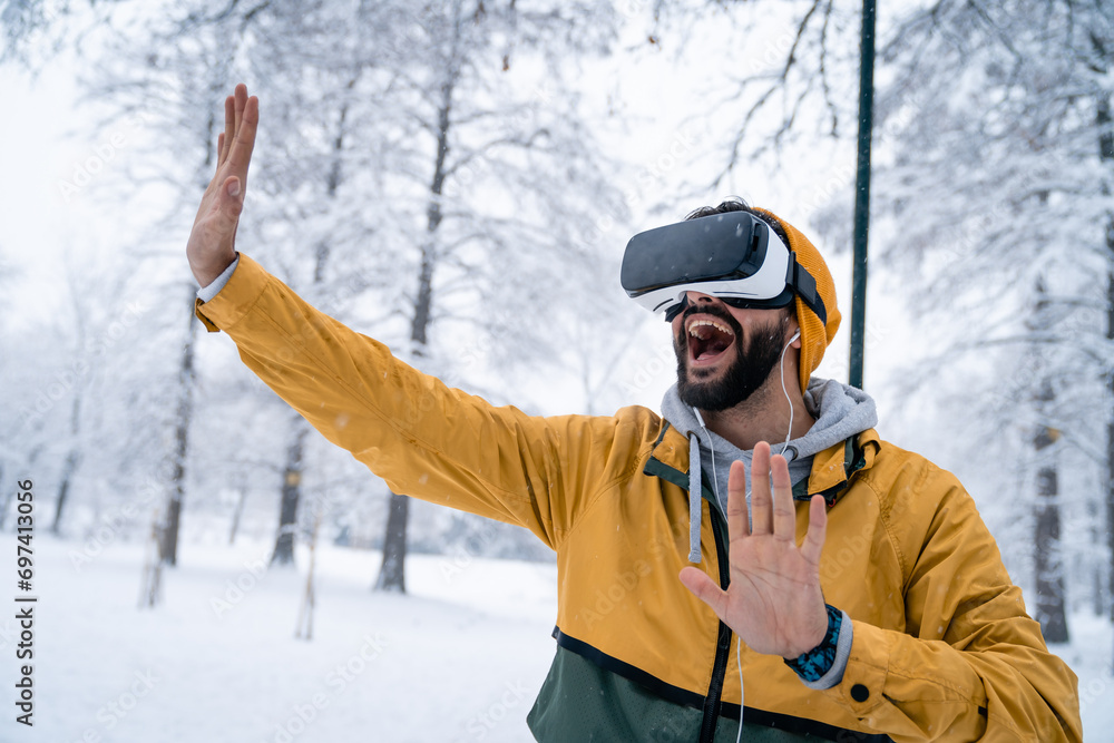Excited man wearing virtual reality glasses gesturing with hands having fun outdoors on snowy day in city park.