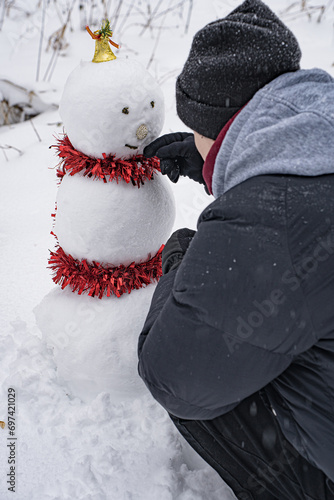 Young guy sculpting and decorating snowman in winter snowy forest