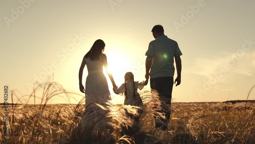 Silhouette of mother with father holding hands of funny daughter jumping over grass. Wife and husband walk looking at daughter strolling and jumping. Mother with father walking at sunset on field photo