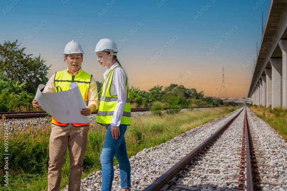 construction crew of a white engineer woman and an Asian man wearing hard hats and reflective vests hold blueprints, watching construction work, inspecting railroad maintenance. construction crew of a white engineer woman and an Asian man wearing hard hats and reflective vests hold blueprints, watching construction work, inspecting railroad maintenance.
