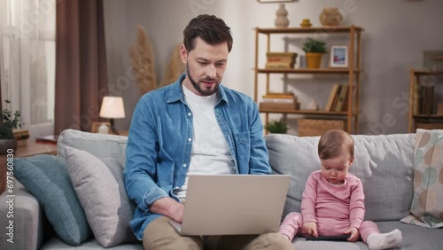 Man taking care of little daughter and working at home. Father babysitting with child. Man raising head and smiling at camera. Kiddo playing with phone. Father and daughter on sofa. photo