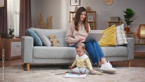 Woman sitting at sofa and holding laptop. Mother working online and taking care of daughter. Baby girl sitting at carpet floor and playing with colored pencils. Mother and toddler at home. photo