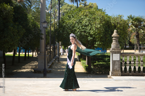 Young, pretty, blonde woman in a green party outfit with sequins, a diamond crown and a beauty pageant winner's sash, twirling so that the air blows her cape. Concept of beauty, fashion, pageant. photo