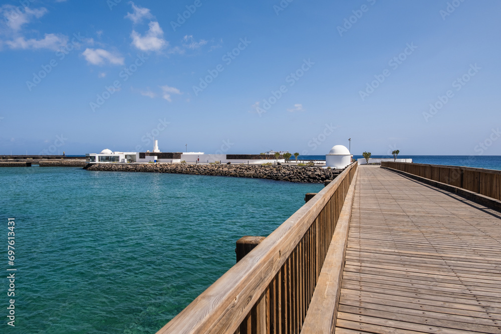 View of the Fermina islet and wooden access bridge. Turquoise blue water. Sky with big white clouds. Seascape. Lanzarote, Canary Islands, Spain. View of the Fermina islet and wooden access bridge. Turquoise blue water. Sky with big white clouds. Seascape. Lanzarote, Canary Islands, Spain.