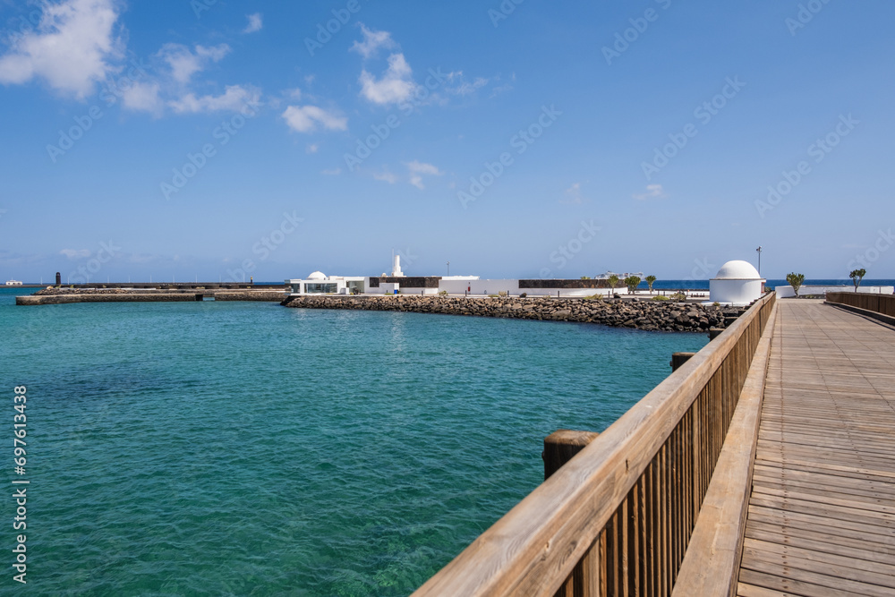 View of the Fermina islet and wooden access bridge. Turquoise blue water. Sky with big white clouds. Seascape. Lanzarote, Canary Islands, Spain.