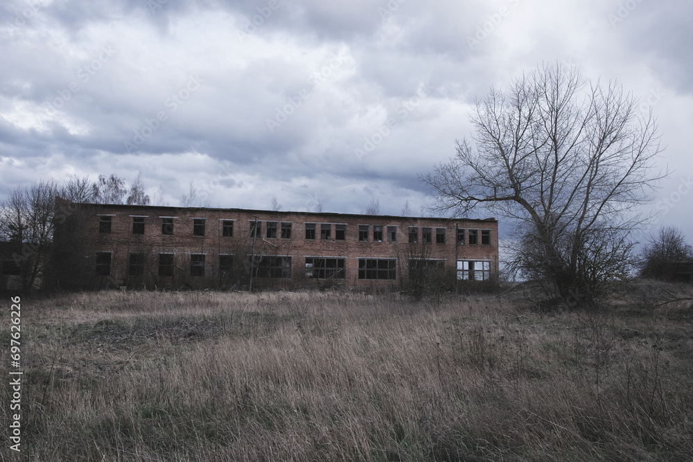 A dilapidated building with broken windows amidst overgrown grass under a cloudy sky.